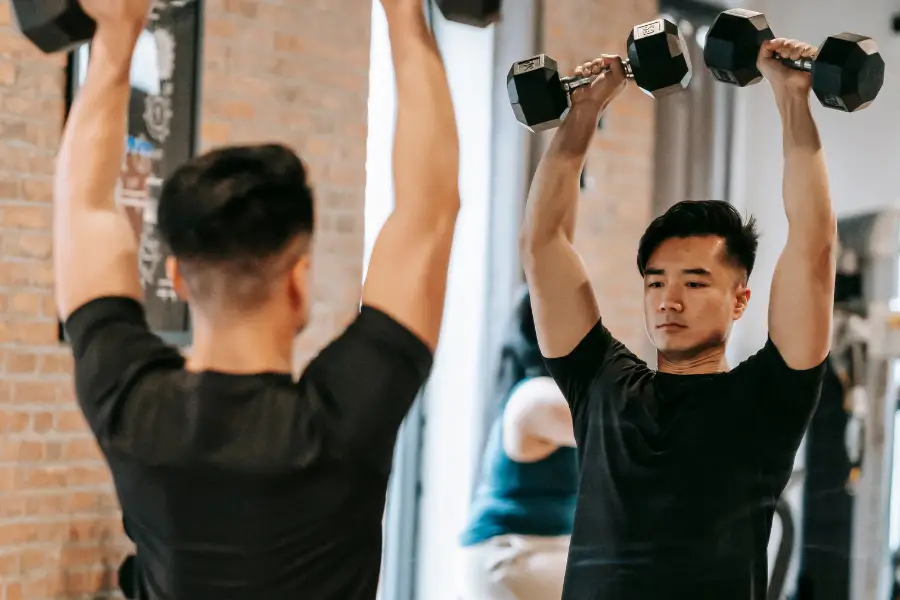 Man lifting dumbbells overhead looking at his reflection in a gym mirror.